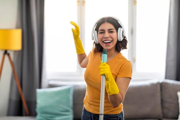 Photo cheerful arab female in wireless headphones singing while making cleaning at home