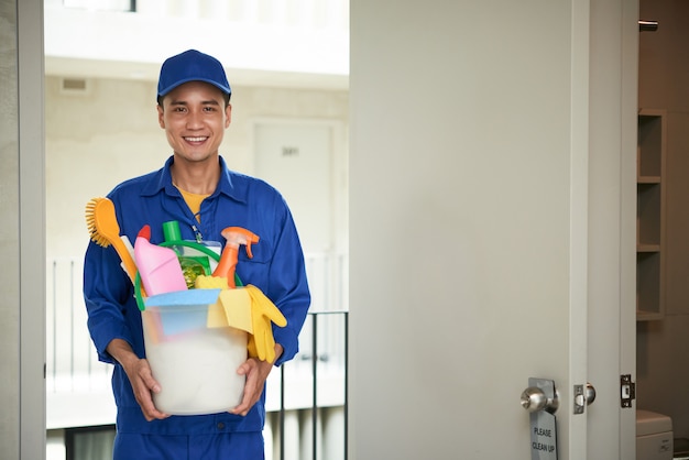 Free photo cheerful asian male janitor walking into hotel room, carrying supplies in bucket