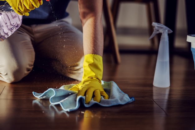 Photo close up of worthy man kneeling, spraying detergent and cleaning parquet at home.