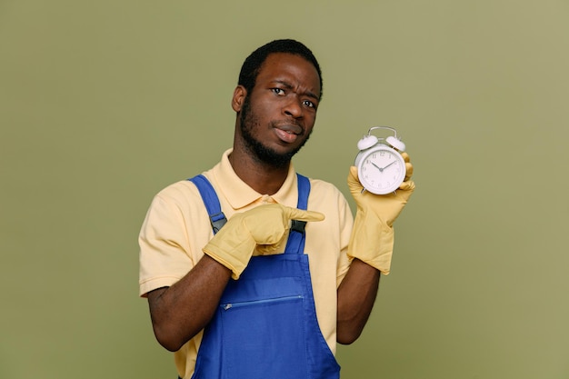 Free photo confident holding and points at alarm clock young africanamerican cleaner male in uniform with gloves isolated on green background