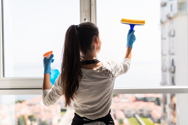 Free photo people, housework and housekeeping concept. happy woman in gloves cleaning window with rag and cleanser spray at home