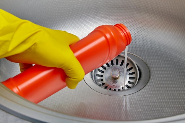 Photo person's hand in a yellow rubber glove pours pipe cleaner down the drain of a metal kitchen sink