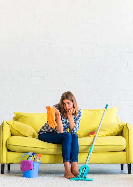 Free photo sad young woman sitting on yellow sofa looking at orange rubber gloves