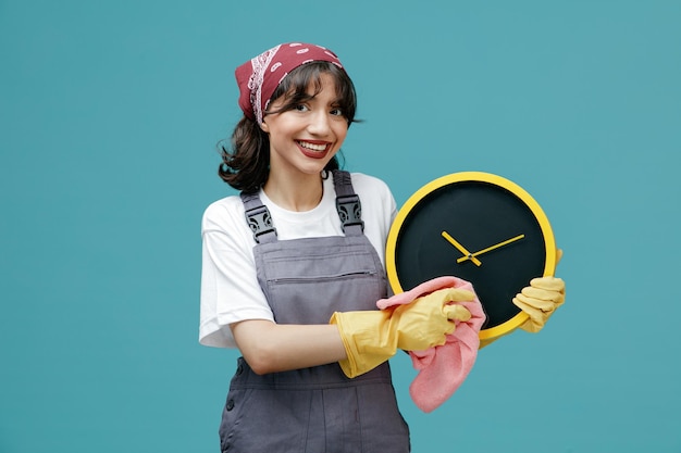 Free photo smiling young female cleaner wearing uniform bandana and rubber gloves holding clock wiping it with duster looking at camera isolated on blue background