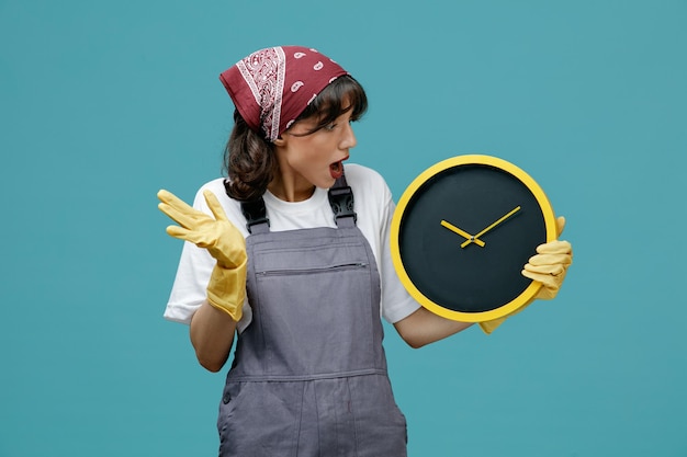 Free photo surprised young female cleaner wearing uniform bandana and rubber gloves holding and looking at clock showing empty hand isolated on blue background