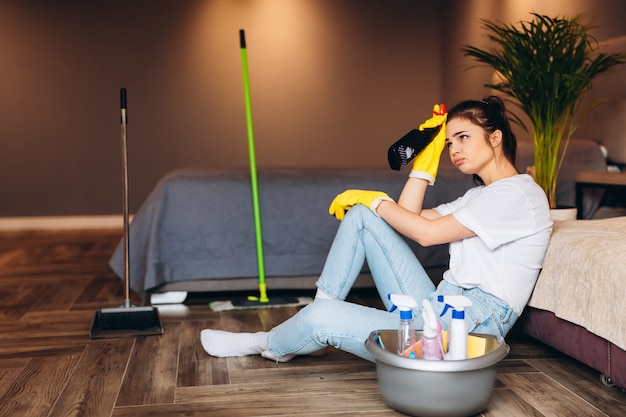 Photo tired woman in white t shirt with dark hair cleaning in yellow rubber gloves for hands protection and bucket with cleaning supplies at home