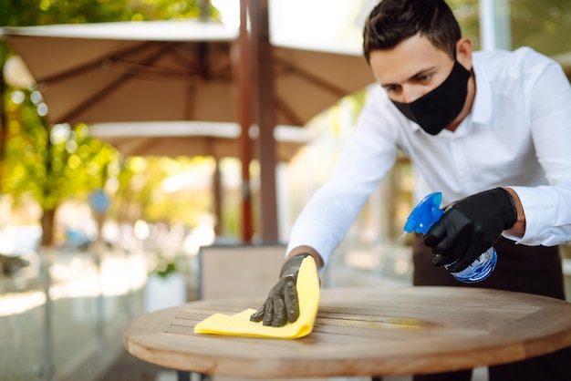 Photo waiter wearing protective face mask and gloves while disinfecting tables at outdoor cafe.