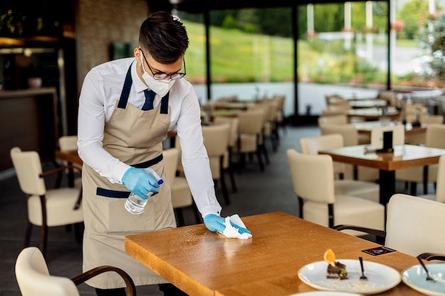 Free photo waiter with protective face mask disinfecting tables in a cafe due to coronavirus epidemic