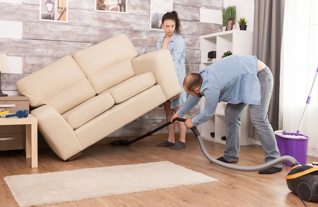 Free photo wife picks up sofa while her husband is cleaning the dust under it with vacuum cleaner