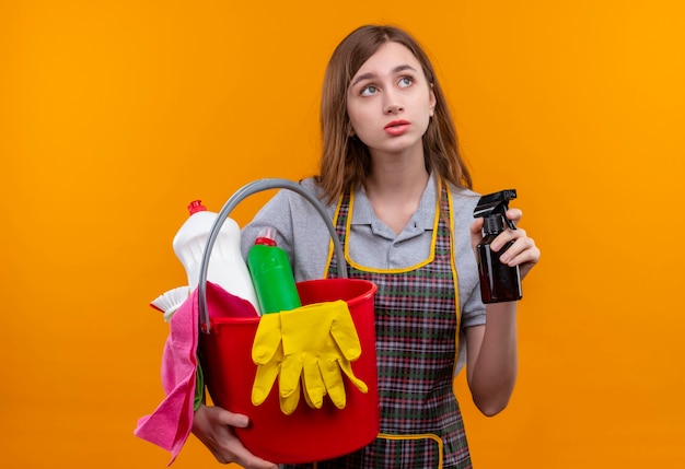 Free photo young beautiful girl in apron holding bucket with cleaning tools and cleaning spray looking aside with sad expression on face