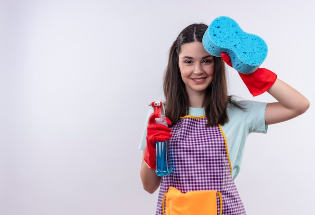 Free photo young beautiful girl in apron and rubber gloves holding cleaning spray showing sponge smiling cheerfully
