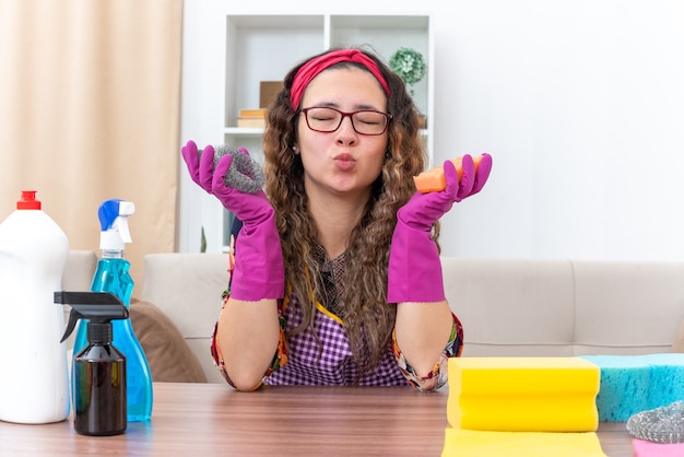 Free photo young woman in rubber gloves happy and positive blowing a kiss sitting at the table with cleaning supplies and tools in light living room